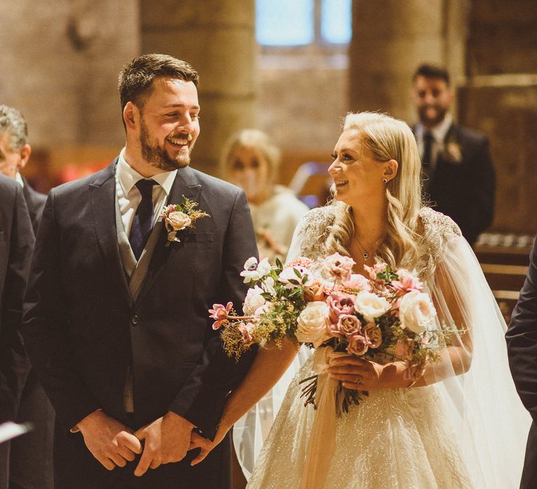 Bride and groom smiling at the altar with bride in a wedding cape holding a blush bouquet 