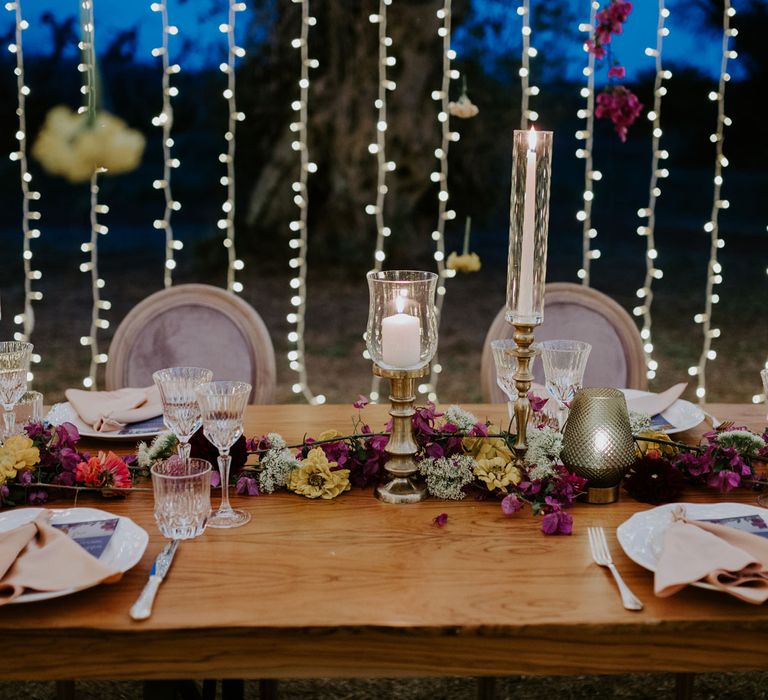 Glass and bronze candle holders, cut crystal glasses and multicoloured floral table garland on rustic wooden table with fairy light backdrop at enchanted forest wedding in Italy