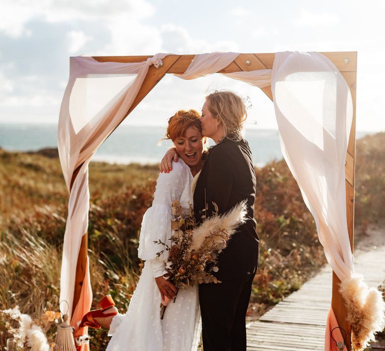 Female same-sex couple under arch on beach for boho elopement