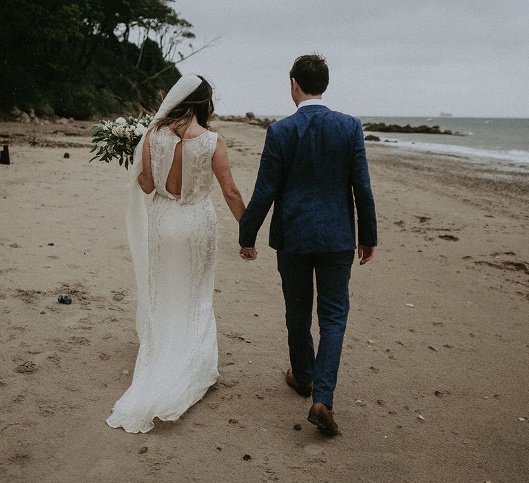 Bride & groom walk hand in hand along the beach on their wedding day