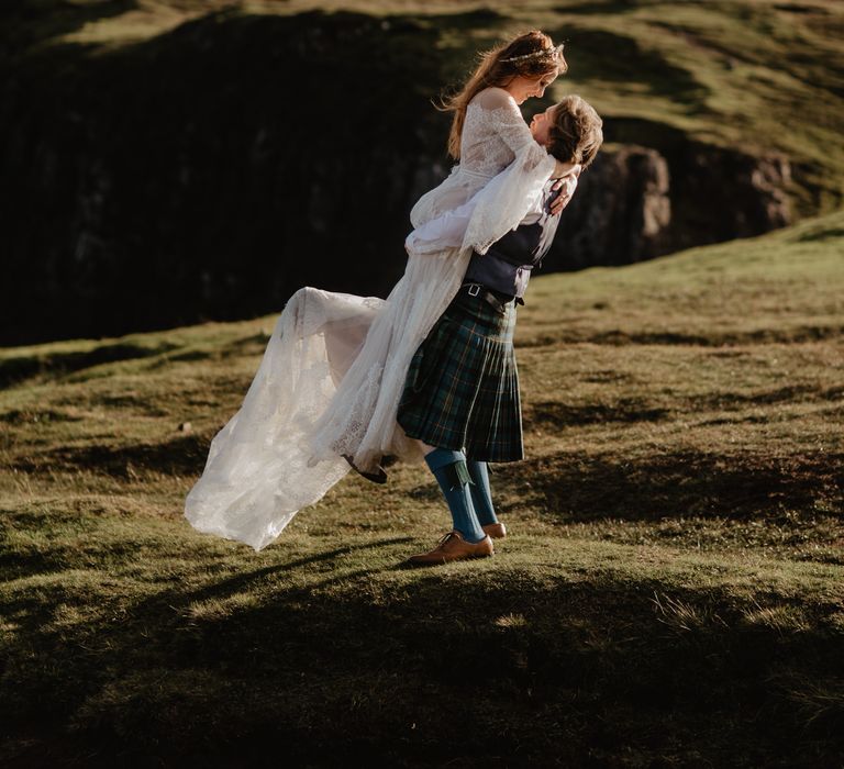 Groom picks up bride on the hillside as the sun shines behind them