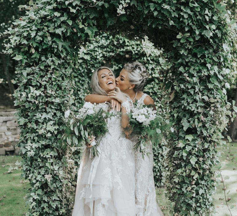 Two brides embrace under ivy covered arches at Crab & Lobster fairytale wedding.