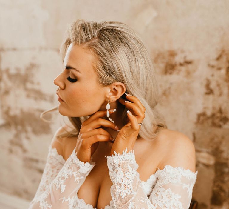 Bride wearing an off the shoulder lace wedding dress fixing her white drop earrings by The Vamoose