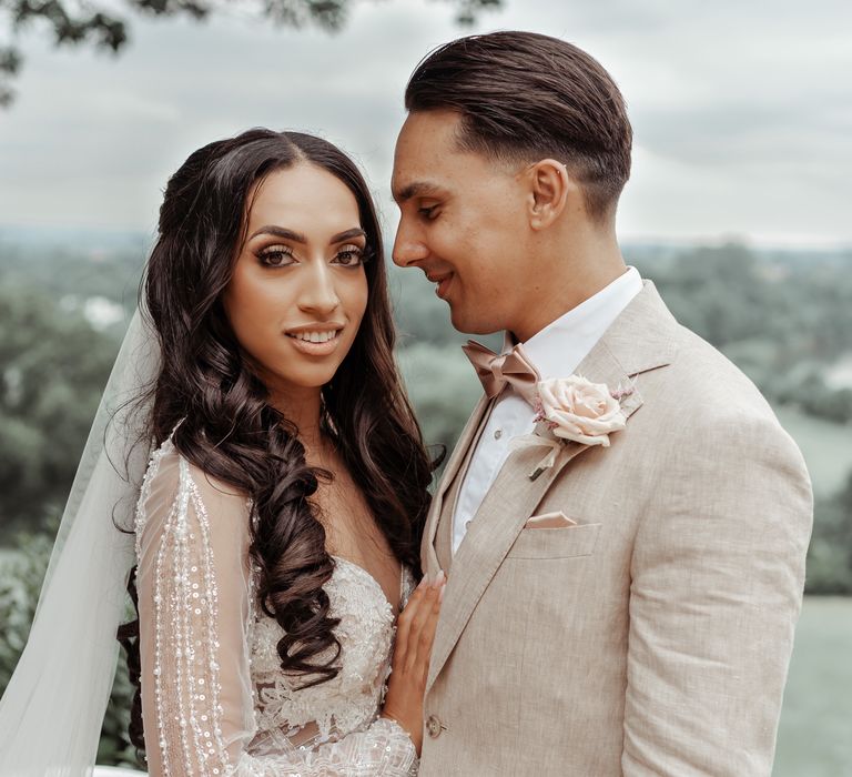 Bride & groom stand outdoors as groom looks lovingly at bride as he wears neutral coloured suit