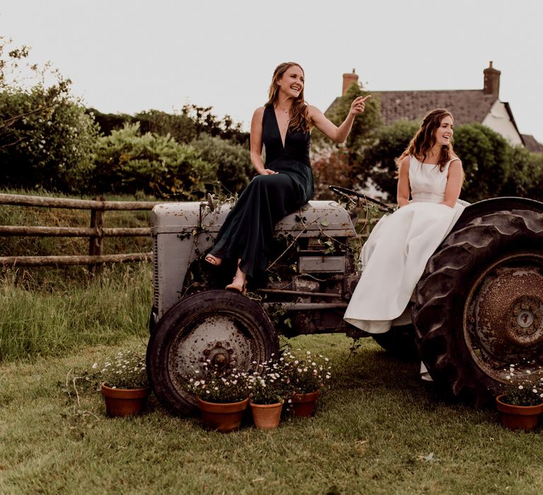 Bride in white Elbeth Gillis wedding gown sits on old tractor with bridesmaid in forest green bridesmaids dress at home farm wedding