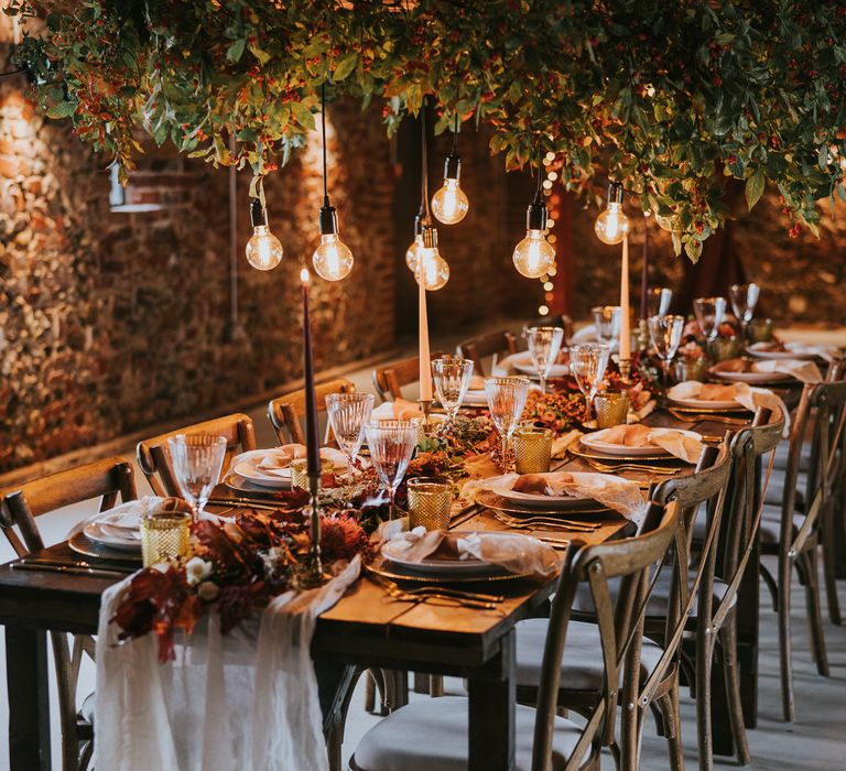 Wooden banquet table with gold and orange tablescape details, underneath a hanging foliage installation with Edison bulbs