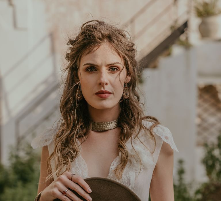 Bride looks intensely toward the camera as she holds her grooms hat and wears her hair in natural curls with minimalistic makeup