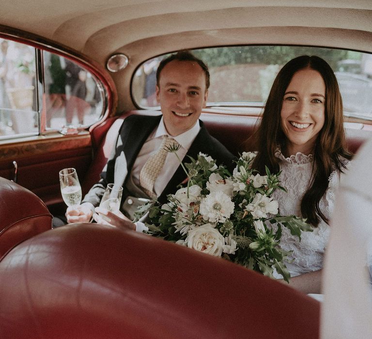 Bride in white lace puffed sleeve dress holding white and green bridal bouquet sits on red leather seats in the back of a classic car with groom in morning coat as he holds two classes of sparkling wine at Surrey summer wedding