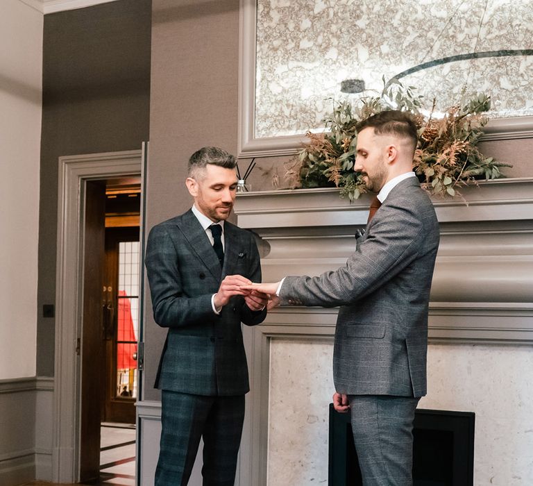 Groom places ring on his husband during wedding ceremony in front of fireplace in the Old Marylebone Town Hall