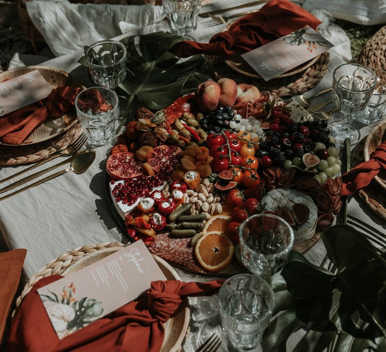 Rustic boho round wedding grazing platter on low white table with palm leaves, boho tableware and low cushions for birds of paradise wedding inspiration