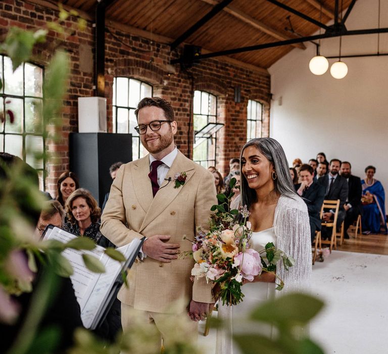 Groom in double breasted linen suit and gold Nike Air Force 1 trainers holds hands with bride in satin Halfpenny London wedding dress and tasselled bridal cape holding multicoloured bridal bouquet during wedding ceremony at Loft Studios London