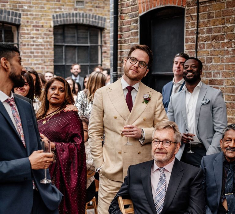 Groom in double breasted linen suit with gold Nike Air Force 1 trainers and burgundy tie holds glass of sparkling wine standing amongst wedding guests listening to speeches outside at Loft Studios London