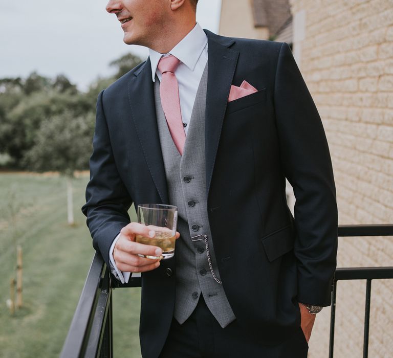 Groom in dark three piece suit with grey waistcoat, pink tie and pink pocket square holding drink whilst he looks out from balcony before Tythe Barn wedding with barn wedding flowers