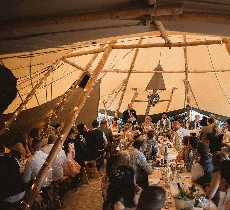 Wedding guests sit in tipi with long wooden tables and fairy lights during wedding breakfast at Inkersll Grange Farm wedding