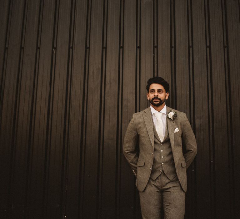 Groom in grey three piece suit with white tie, pocket square and floral buttonhole leans against barn wall at Inkersall Grange Farm wedding 