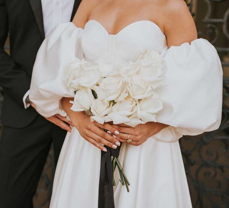 Bride in a sweetheart neckline wedding dress with detachable puff sleeves holding an all white rose wedding bouquet showing off her iridescent nail polish 