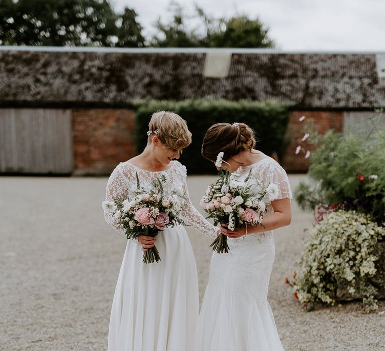 Two brides meeting at their first look in lace and embellished wedding dresses holding pink and white wedding bouquets 