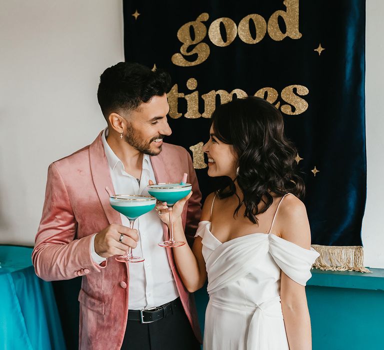 Groom in pink velvet jacket and bride in a cold shoulder wedding dress sipping cocktails in front of a 'let the good times roll' fabric banner 