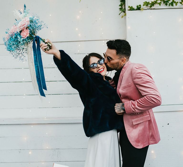 Groom in a pink velvet jacket kissing his brides cheek in a navy faux fur coat as she holds her pink and teal wedding bouquet in the air 