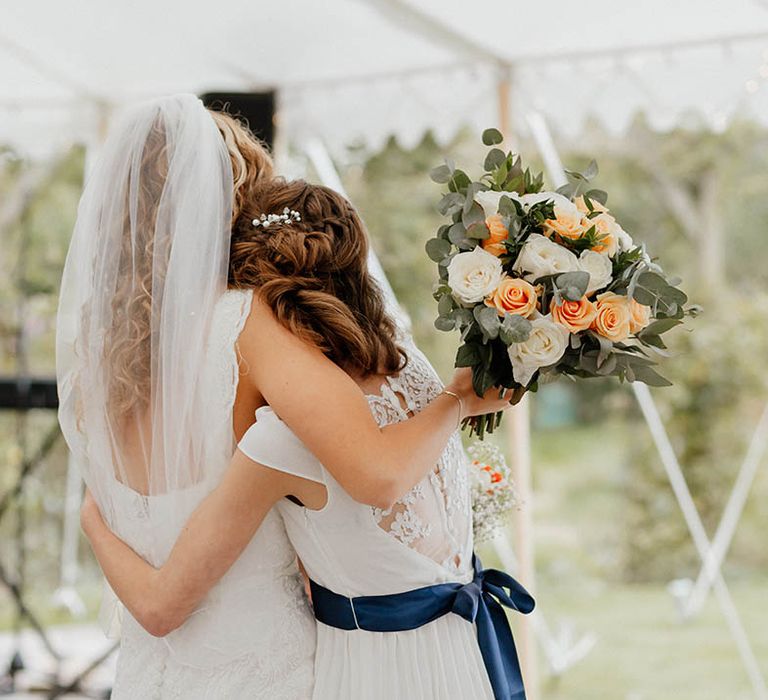 Bride cuddles bridesmaid on her wedding day as they look out toward garden in marquee