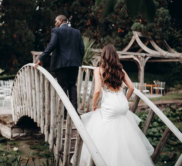 Bride in chiffon bottom dress with shaped plunge wedding dress walks over countryside bridge with Groom in dark fitted suit