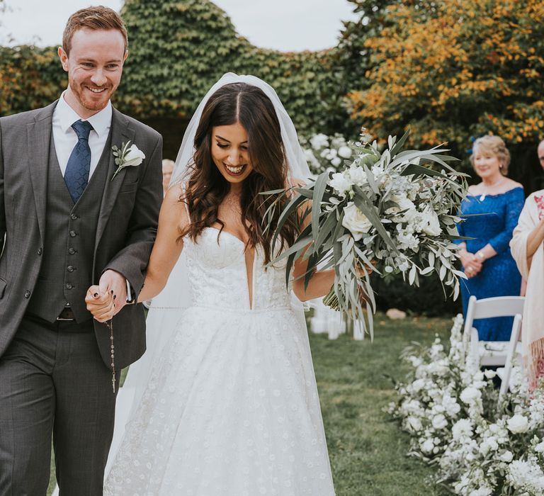 Bride in white cami strap applique Pronovias wedding dress and veil holding green and white rose bridal bouquet walks down the aisle holding hands with groom in grey three piece suits with blue tie and white rose buttonhole during outdoor wedding ceremony in Buckinghamshire