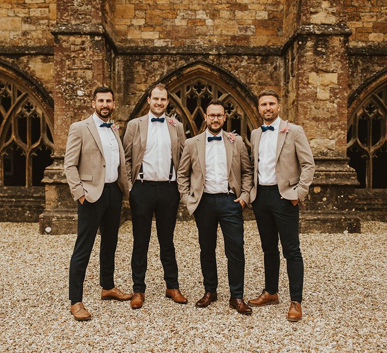 Groom stands with his groomsmen who wear light coloured jackets and bow ties with pastel pink buttonhole florals