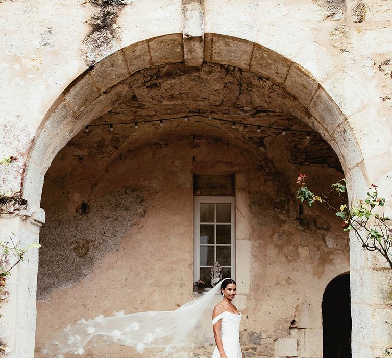 Brides veil blows in the wind as she stands under archway outdoors 