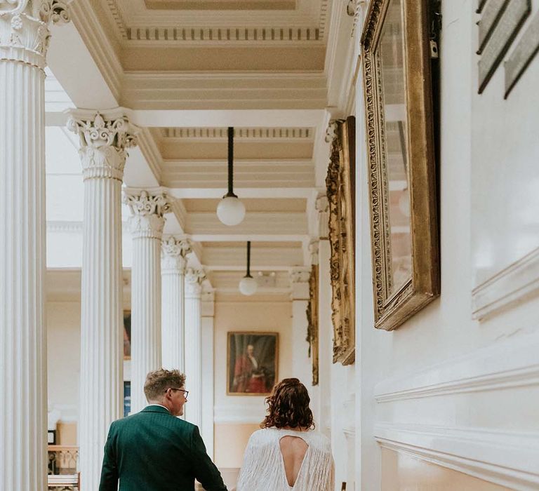 Bride & groom walk away together whilst holding hands and bride wears tassel wedding cape 