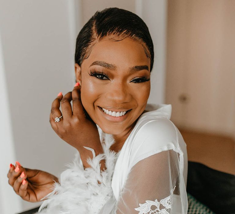 Bride puts earrings in on the morning of her wedding as she wears white dressing gown complete with feathered sleeves