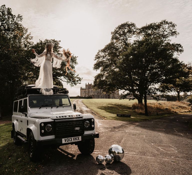 Bride in lace boho two piece wedding dress and bridal hat holding boho bouquet stands on top of Land Rover defender in the grounds of Kinmount House in Scotland