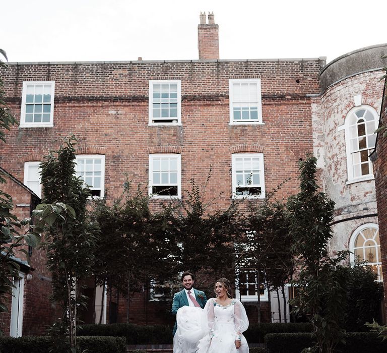 Bride in long sleeve wedding dress and white trainers walks through gardens whilst groom in teal suit holds her train