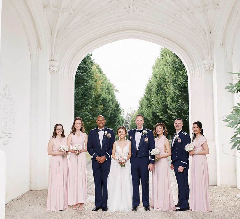 Bride & groom stand with their bridesmaids and groomsmen outdoors under large archway 