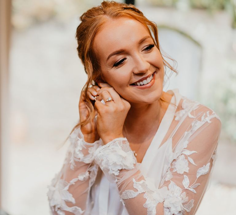 Bride with curled updo waiting mesh and lace bridal robe puts in earrings before her wedding