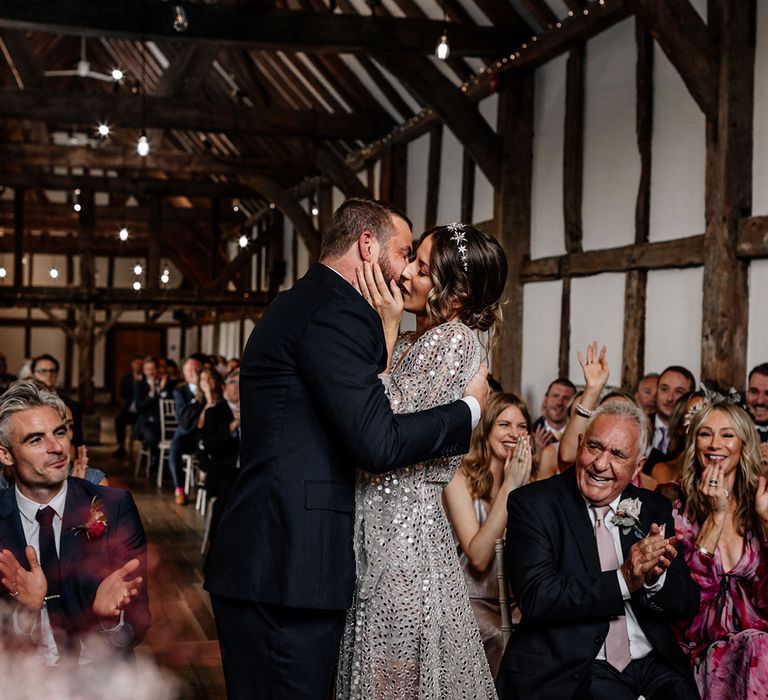 Bride in a silver sequin wedding dress and groom in a navy suit kissing at their Tithe Barn, Loseley House wedding ceremony 