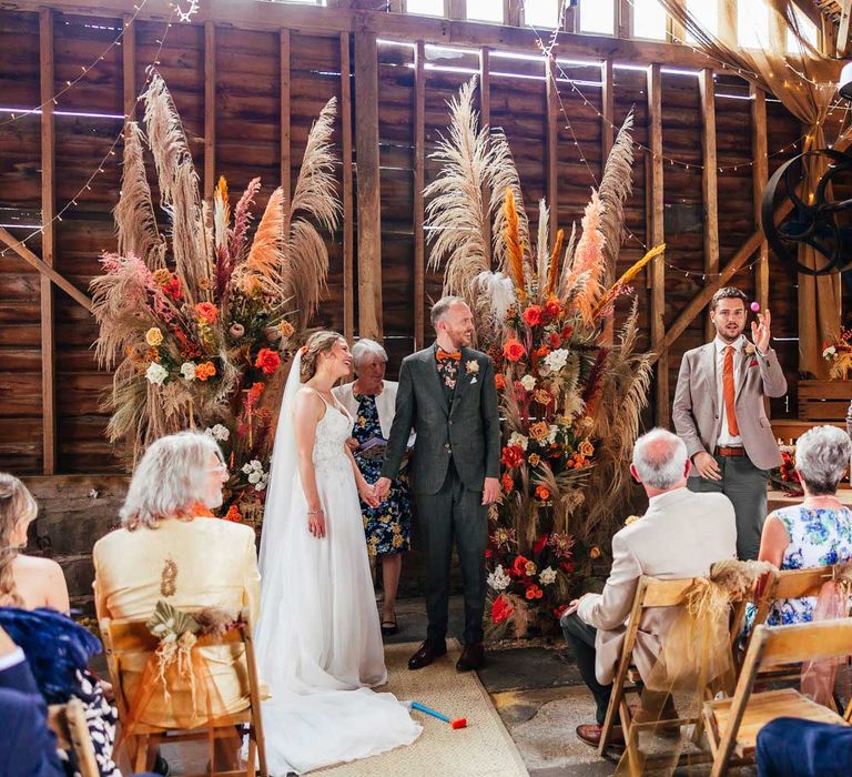 Bride in lace wedding dress and veil holds hands with groom in green suit and patterned shirt with orange bow tie as they watch groomsman in grey suit and orange tie throw ping pong ball during festival style wedding ceremony