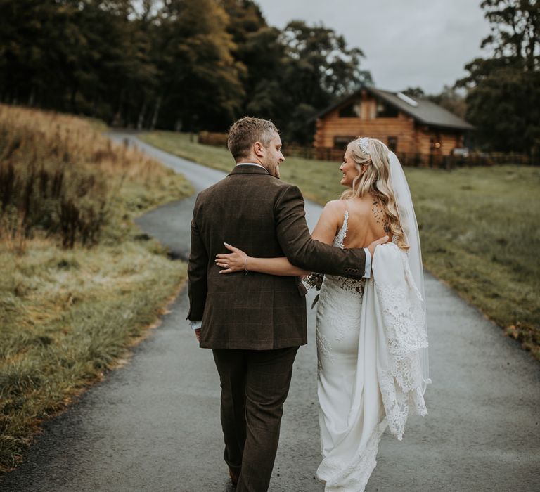 Bride in low back lace wedding dress with button detailing and train walks arm in arm with groom in brown suit as they walk along path at the Hidden River Cabins