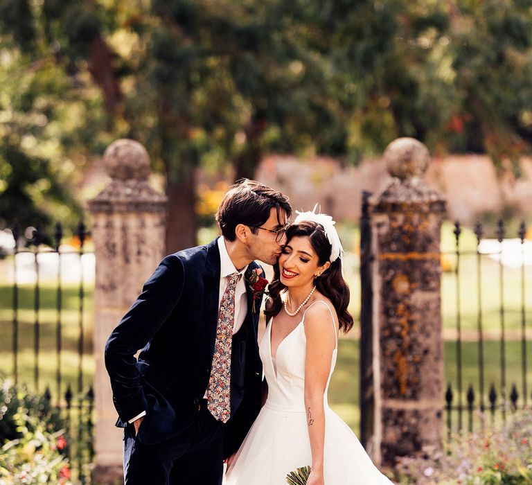 Bride in layered tulle wedding Dres with feather headband and pink and red bridal bouquet stands with groom in dark suit and floral tie for Iranian inspired wedding in the Cotswolds