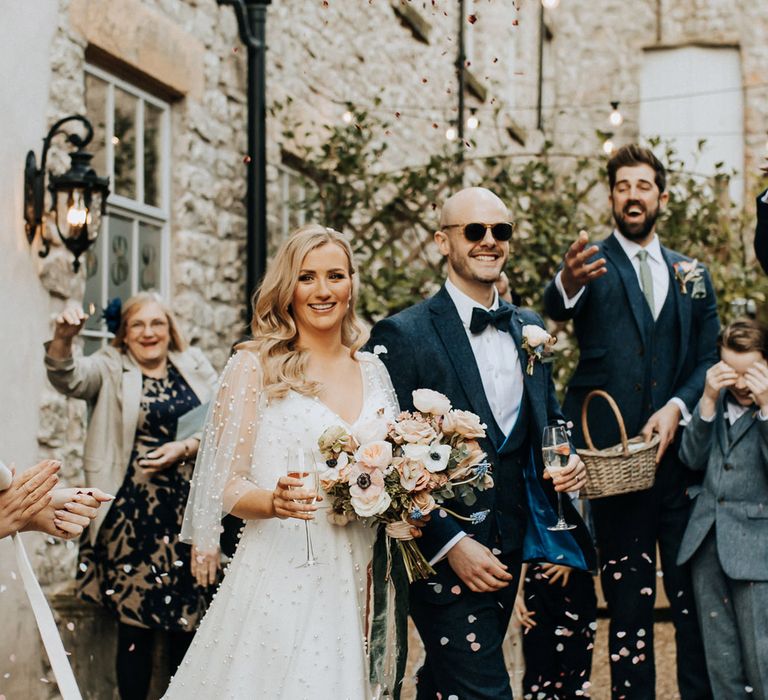 Bride in pearl wedding dress with sleeves holding pastel wedding bouquet and glass of sparkling wine walks out of ceremony with groom navy tweed wedding suit and sunglasses as guests throw confetti