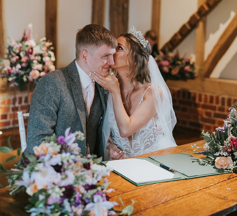 Bride and groom in grey tweed suit sign the registry in rustic wedding venue and large flower displays