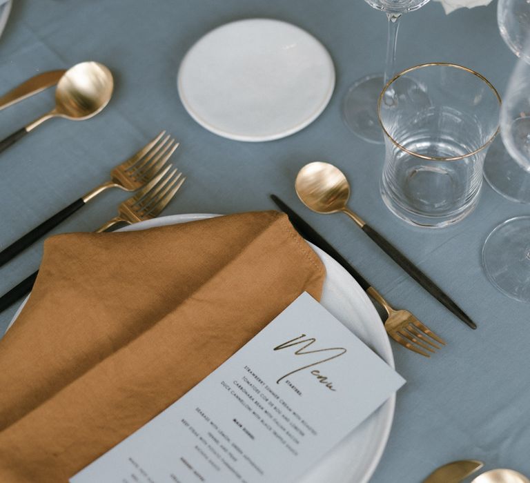 Tablescape with white plate, grey tablecloth, toffee coloured napkin, gold cutlery and light coloured flower arrangement 