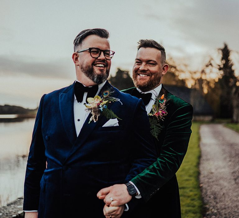 Grooms in colourful velvet suit jackets smile and laugh together as they pose by a lake at Castle Leslie Estate wedding