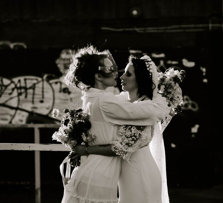 Two brides embracing in long and short wedding dress with a birdcage veil and long embellished veil 