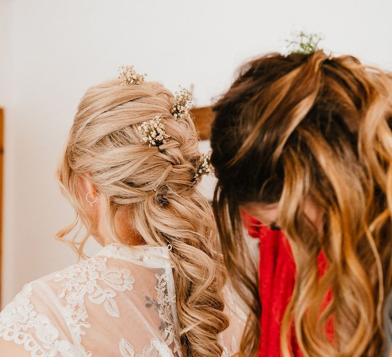 Bride with blonde hair styled into a delicate braid with gypsophila hair pins is buttoned into her wedding dress by friend