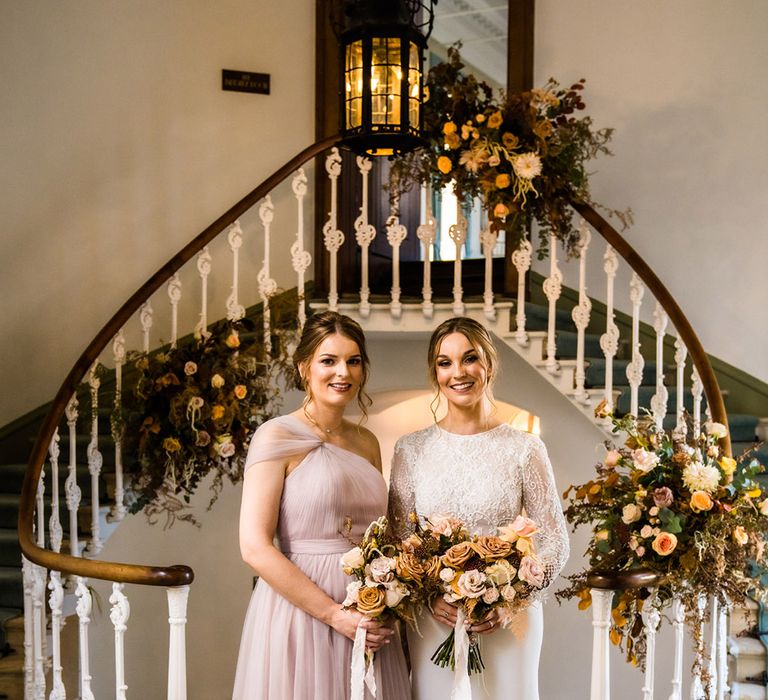 Bride in lace top long sleeve fitted wedding dress with bridesmaid in pale pink wedding dress with tulle skirt and one shoulder detail 