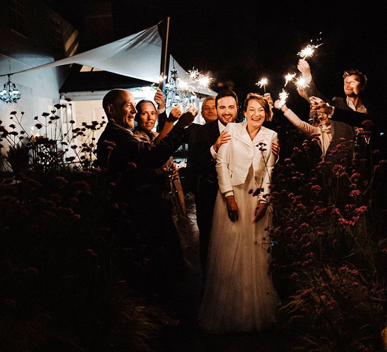 Groom leads the bride wearing white leather cover-up jacket through guests waving sparklers