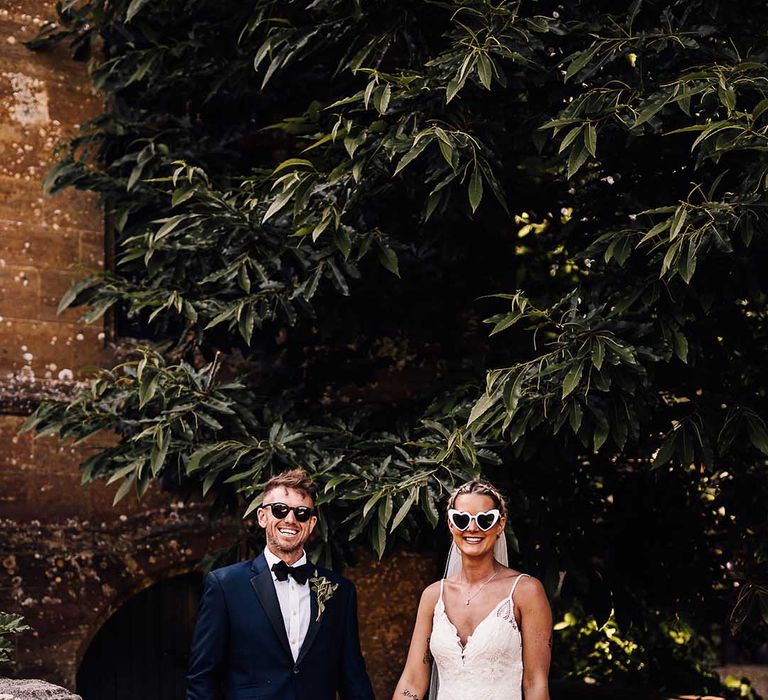 Groom in blue tuxedo and black sunglasses with bride in lace wedding dress and retro heart sunglasses 