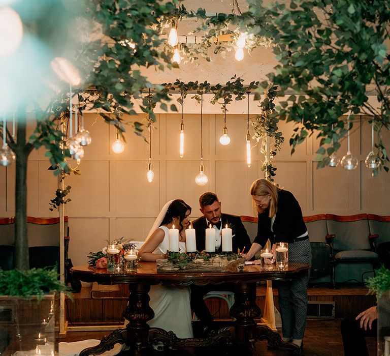 Bride and groom sign their wedding register with exposed lightbulbs and lit candles 