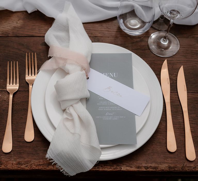 Place setting with white tableware, linen napkin, gold cutlery and grey menu card 