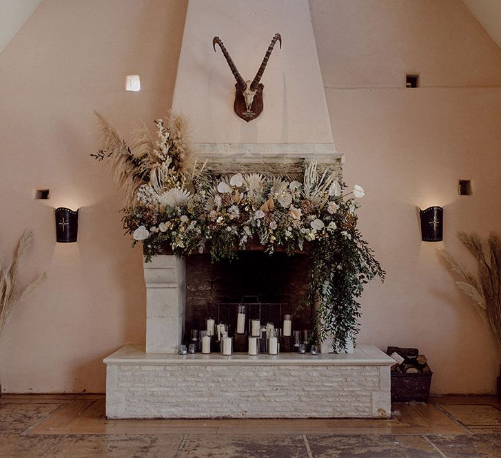 White fireplace surrounded by dried florals and green foliage and white pillar candles  